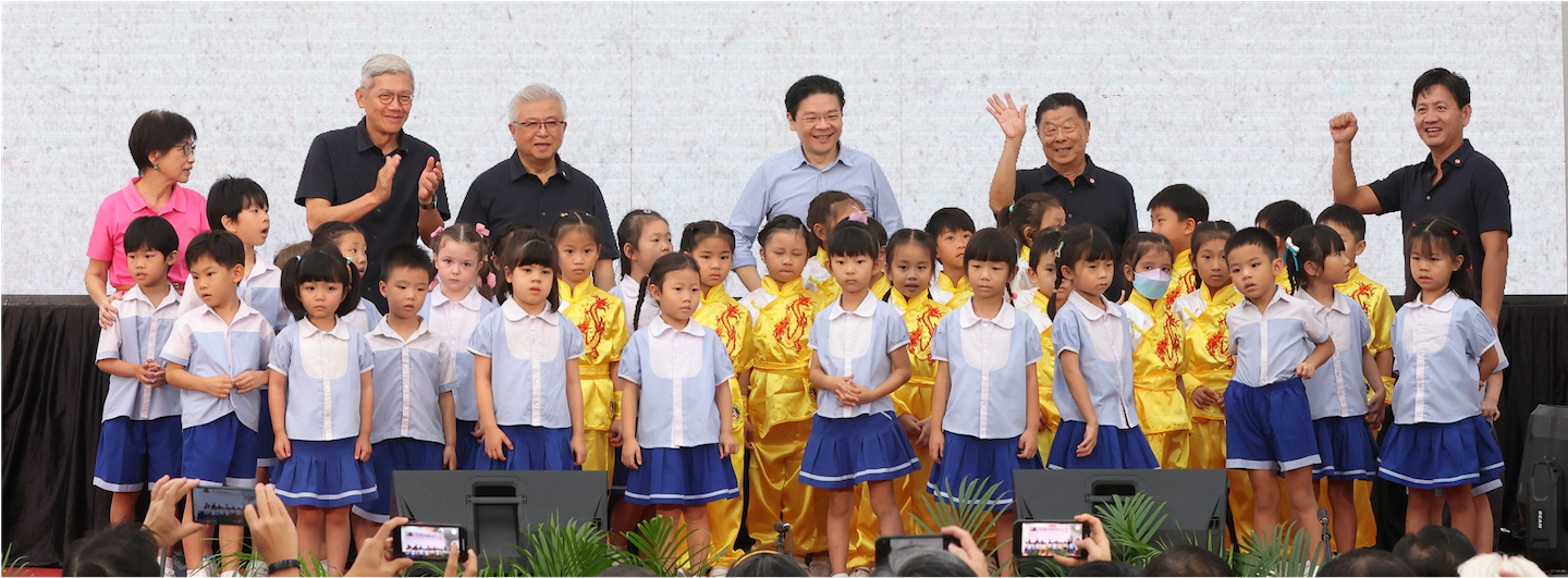 Adults and children on stage, some children wear yellow dragon patterned outfits, others blue and white.